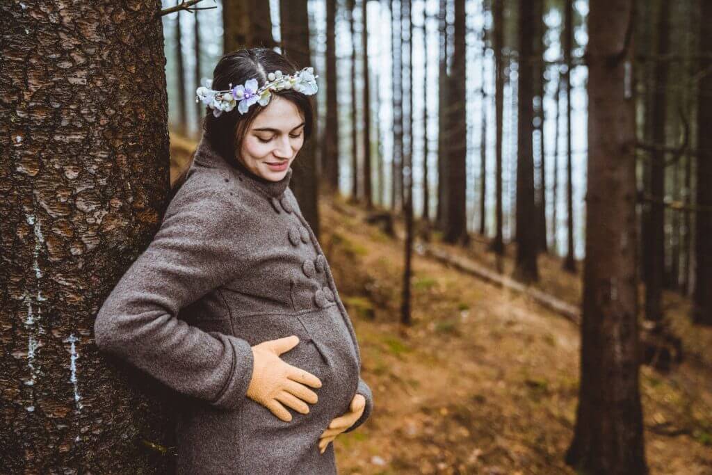 Schwangere lehnt an Baum und blickt auf Babybauch