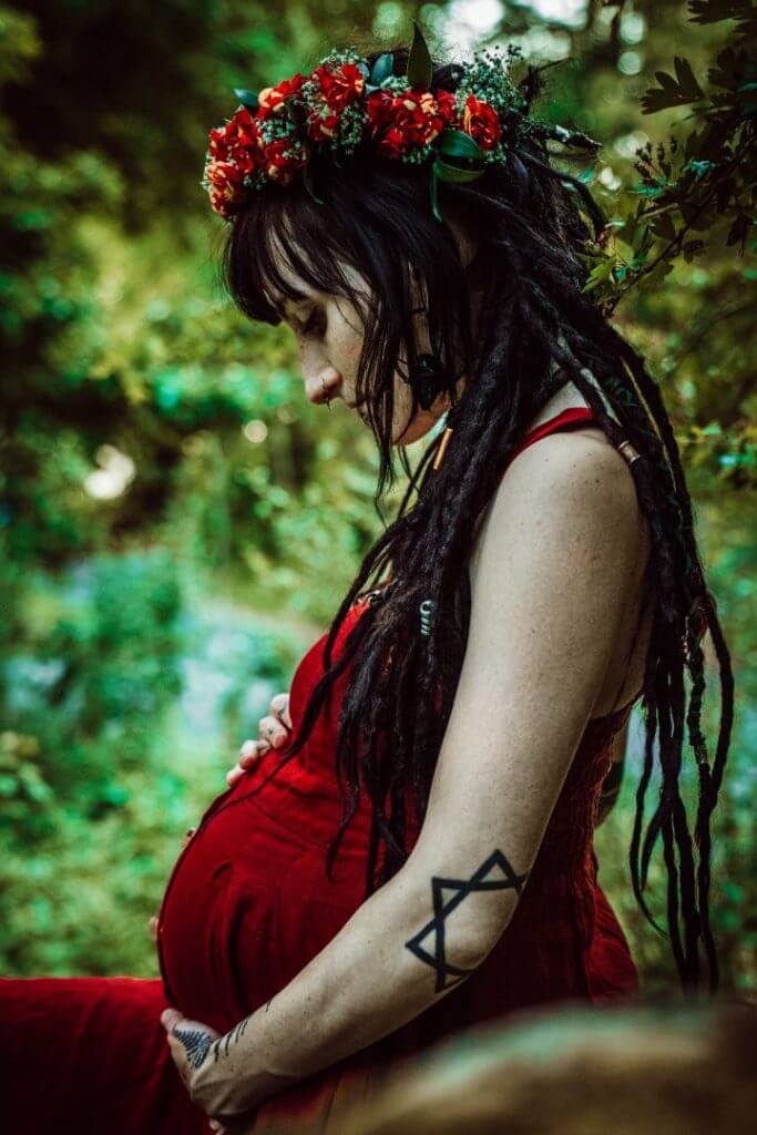 Schwangere sitzt auf Ast und schaut auf Babybauch in rotem Kleid