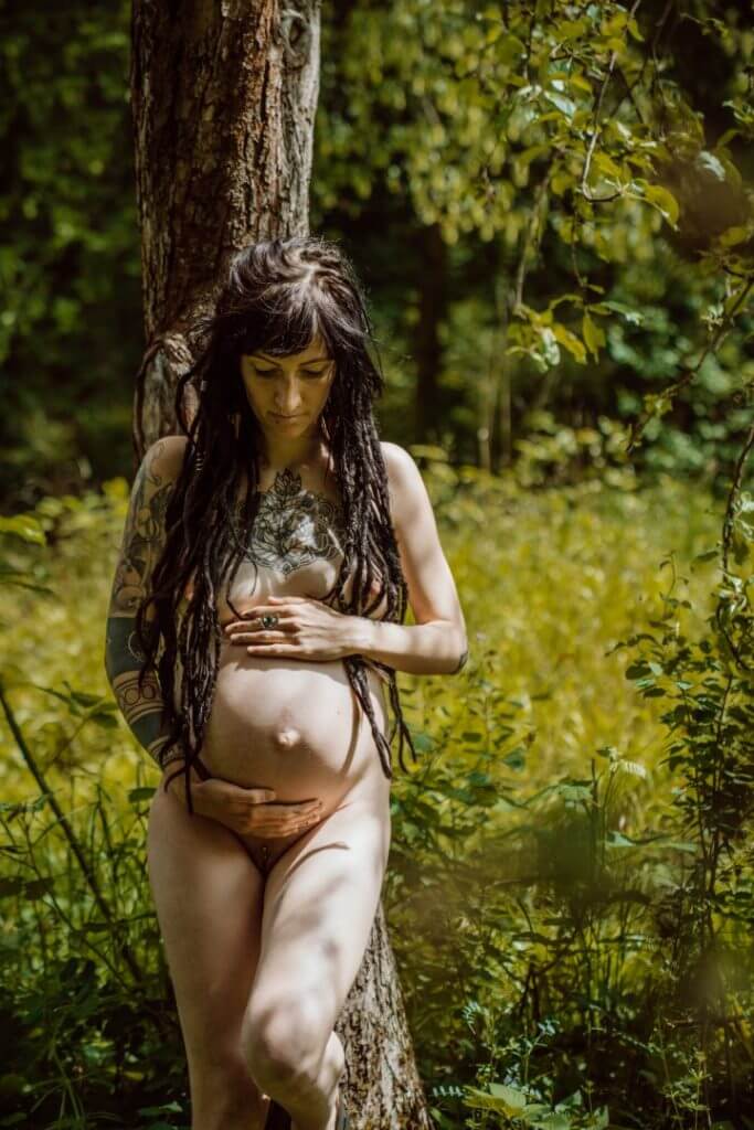 Schwangere lehnt nackt am Baum, hält Babybauch