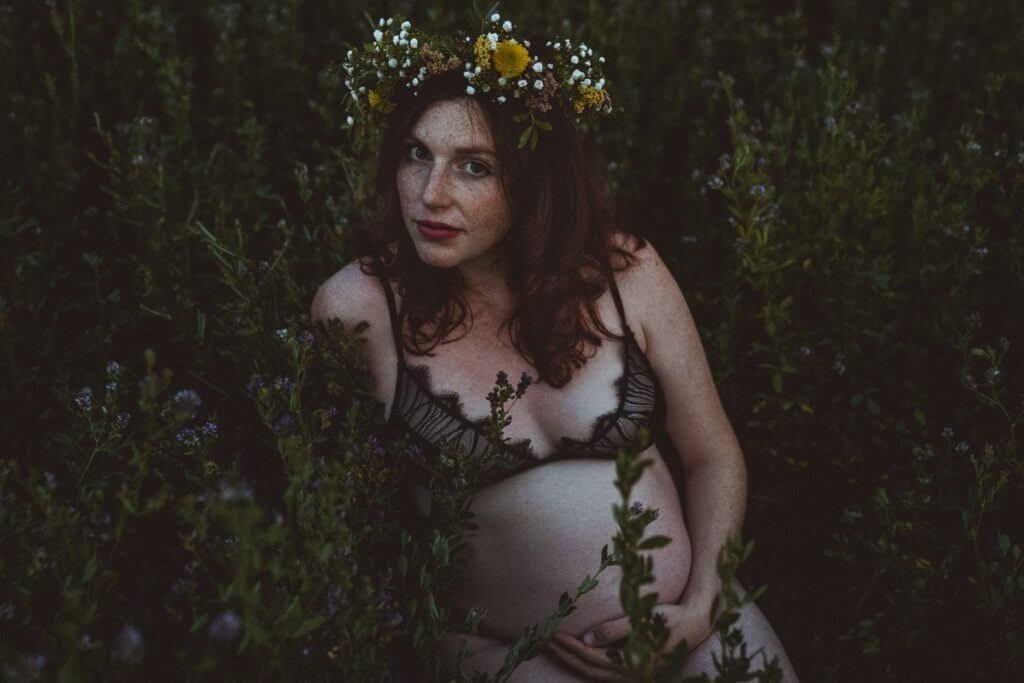 Schwangere mit Blumenkranz sitzt in Unterwäsche auf Wiese