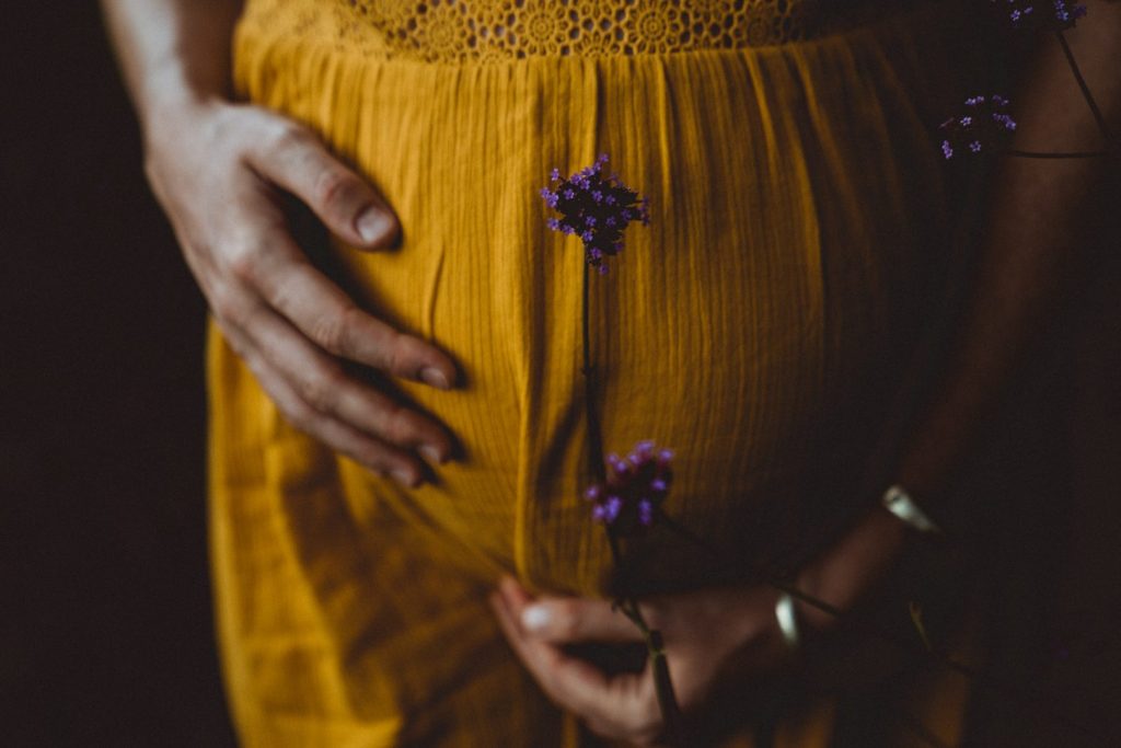Schwangere hält Babybauch mit lila Blüten in gelbem Kleid