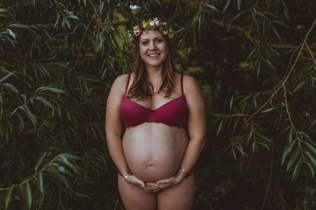 Schwangere steht in Unterwäsche in Weide und lächelt in Kamera