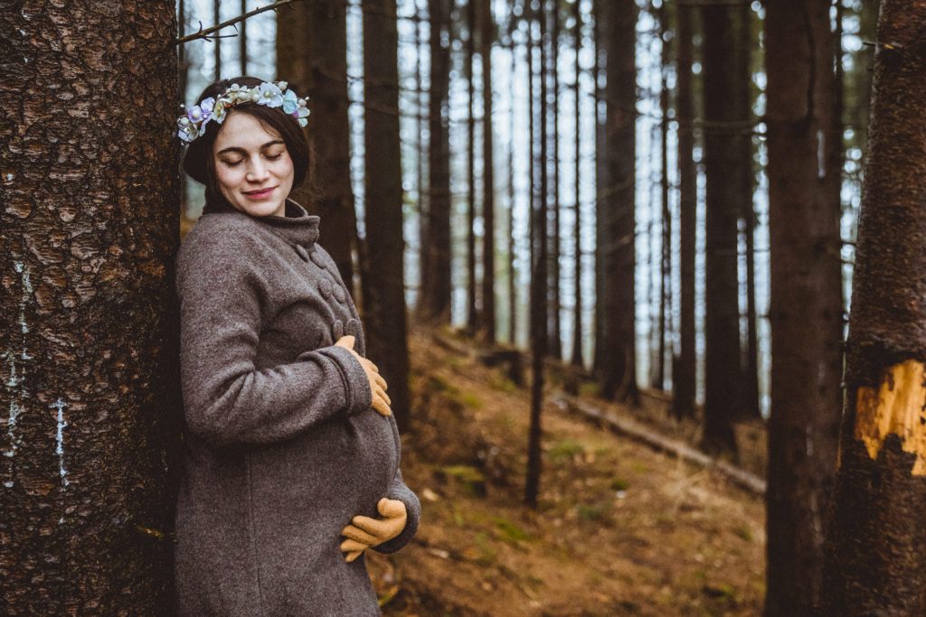 Schwangere in grauem Wintermantel lehnt an Baum