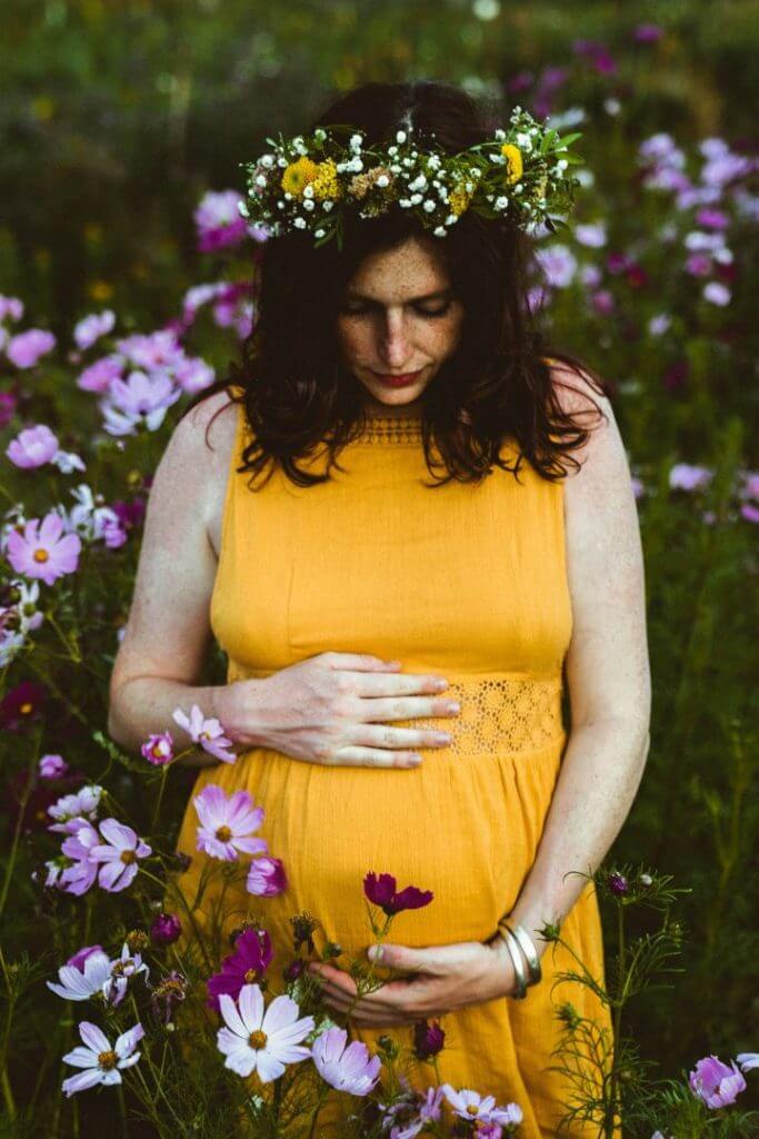 Schwangere trägt Blumenkranz und blickt auf Babybauch in gelbem Kleid