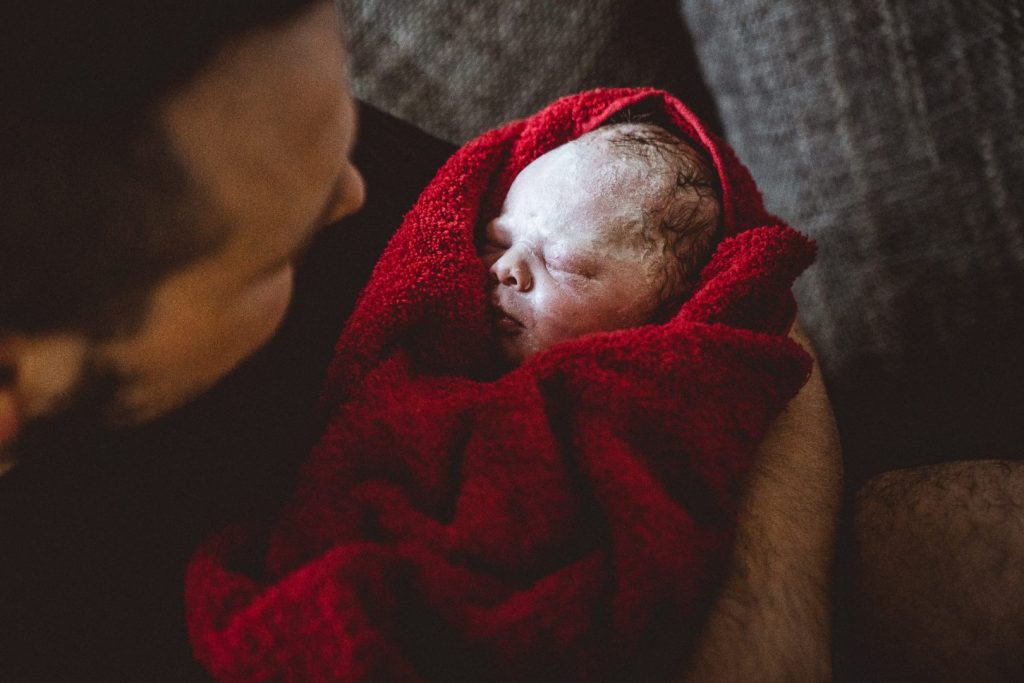 Vater blickt auf Neugeborenes, welches in rotes Handtuch gewickelt ist