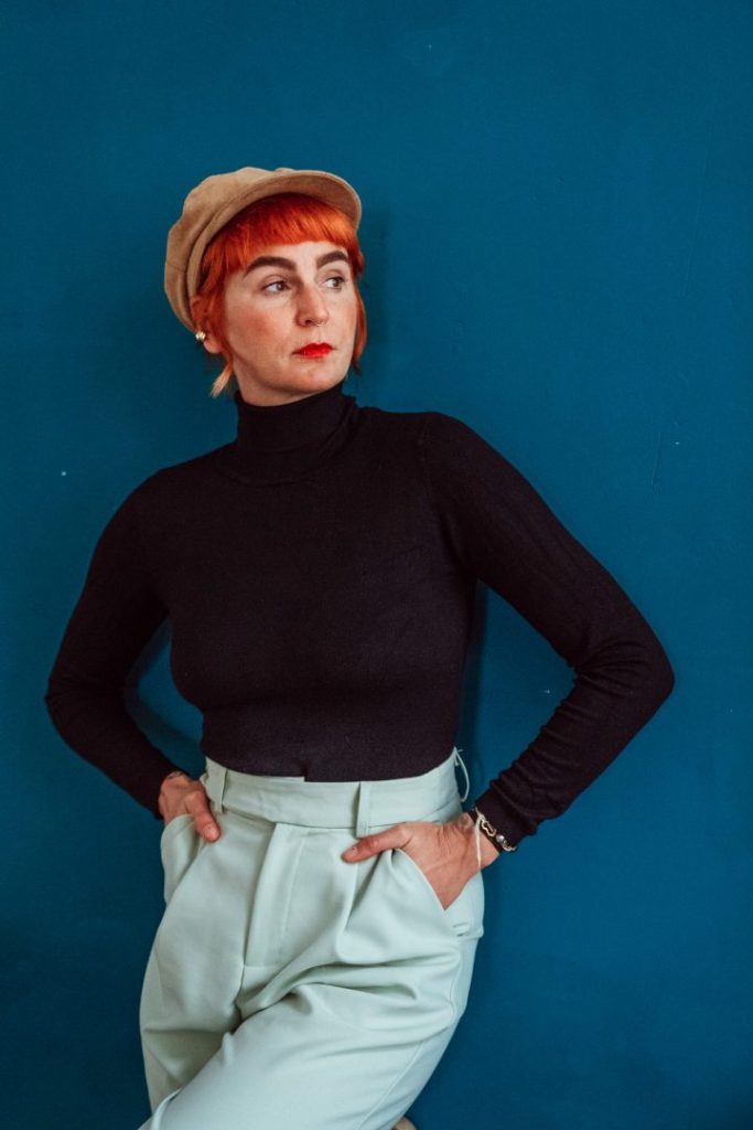 Frau lehnt vor blauer Wand und fässt in Hosentaschen