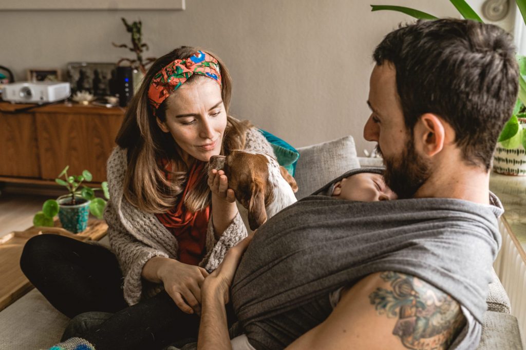 Vater trägt Baby in Tragetuch und Mutter kuschelt Hund