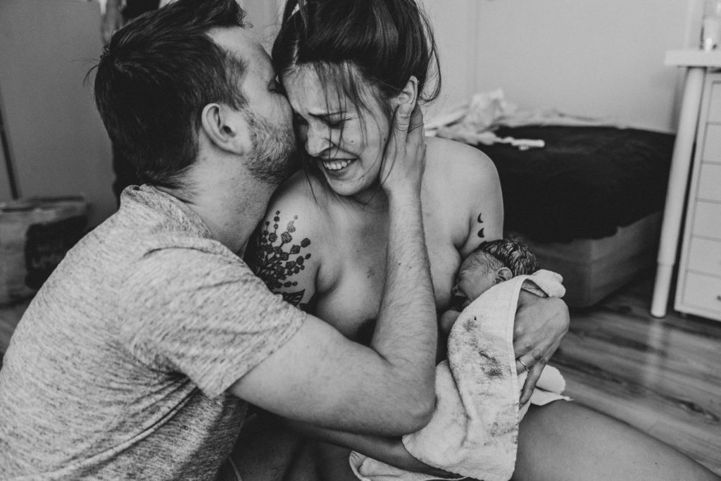 Mutter hält Baby nach Geburt in Arm und wird von Partner geküsst