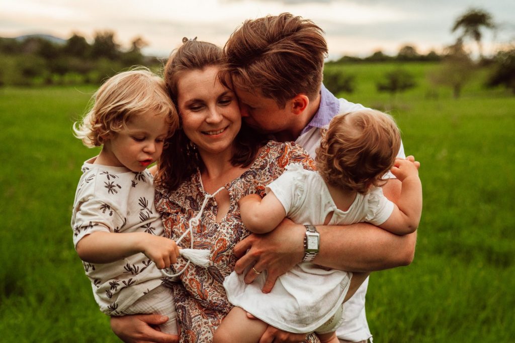 Mama hält Kinder auf dem Arm und wird von Partner geküsst